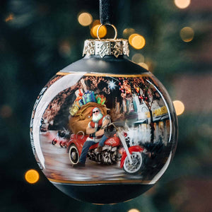 Decorative Christmas ornament with Santa Claus on a motorcycle in a snowy village scene.