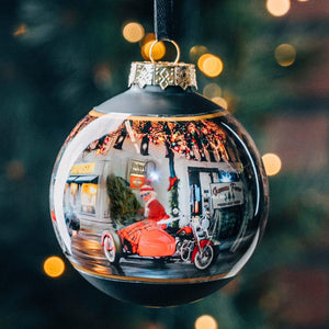 Decorative Christmas ornament with Santa Claus on a motorcycle inside, against a blurred festive background.