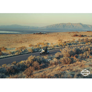 Motorcycle on a desert road with mountains in the background, featuring a Harley-Davidson logo.