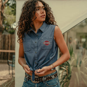Woman wearing a sleeveless denim shirt with a logo, standing outdoors.