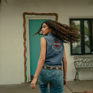 Woman wearing a denim vest with a logo, standing in front of a building with a teal door.