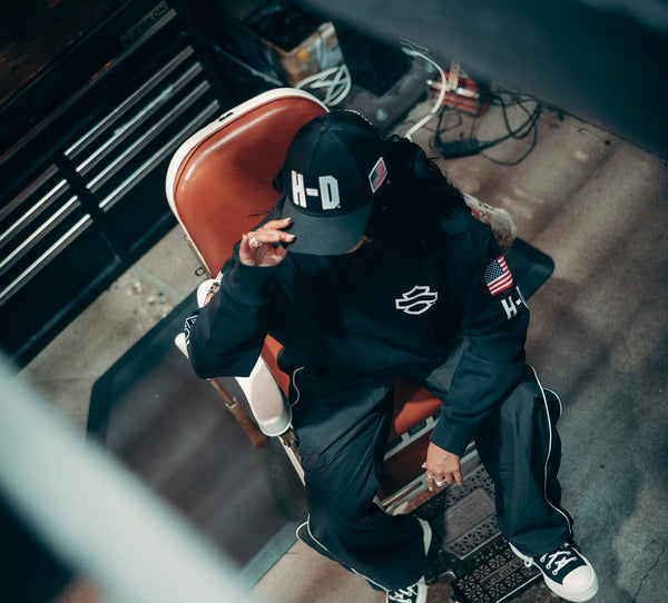 Person wearing a black 'H-D' cap and jacket sitting on a motorcycle in a garage.