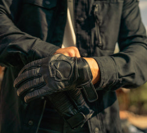 Person wearing black gloves with a logo, holding hands together, against a blurred background.