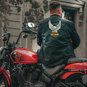 A man wearing a black leather Harley-Davidson jacket with a large eagle and shield patch on the back, sitting on a red cruiser motorcycle in front of a building.