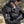 A woman wearing a black leather motorcycle jacket with a white and black 'Harley-Davidson' skull patch on the sleeve, black leather gloves, and a helmet in an outdoor setting.