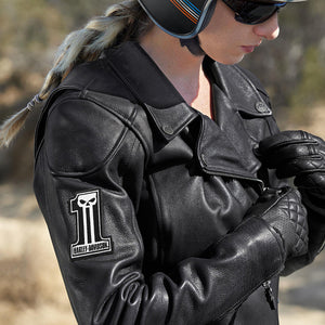 A woman wearing a black leather motorcycle jacket with a white and black 'Harley-Davidson' skull patch on the sleeve, black leather gloves, and a helmet in an outdoor setting.