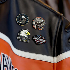 Four Harley-Davidson themed enamel pins displayed on a black leather motorcycle jacket with orange and white racing stripes.