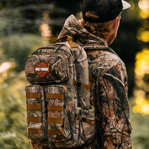 A man wearing a camouflage hunting jacket and cap, shown from behind in a forest, carrying a large camouflage backpack with tan straps and a Harley-Davidson patch on the front.
