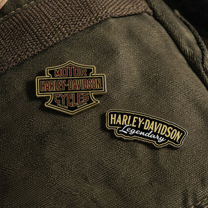 Two Harley-Davidson enamel lapel pins displayed on a dark olive green canvas fabric background. One pin is the classic 'Motor Cycles' shield logo, and the other is a 'Harley-Davidson Legendary' script pin.