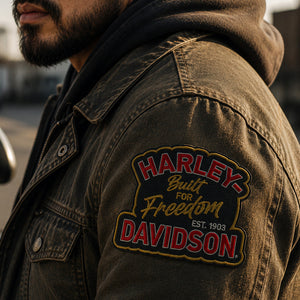 Close-up of a man wearing a dark brown denim jacket with a large embroidered Harley-Davidson patch on the sleeve, set against an outdoor urban background.