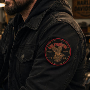 A close-up of a man wearing a black denim jacket with a circular Harley-Davidson embroidered patch on the left sleeve, set in a dimly lit garage environment.