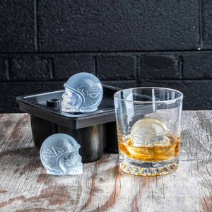 A skull-shaped clear ice cube mold shown with a black mold tray, one molded ice cube, and a whiskey glass containing a large skull-shaped ice cube, set on a rustic wooden surface against a dark brick wall.