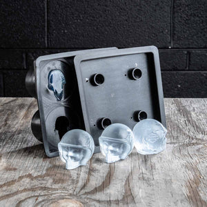 A dark gray silicone mold set shown on a rustic wooden surface with three clear, skull-shaped ice cubes in front. The mold features a square tray design with circular holes and a separate piece with a skull cavity.