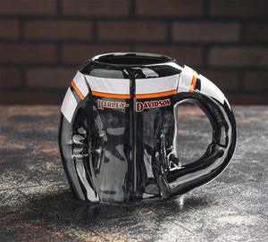 A glossy black ceramic mug with a Harley-Davidson logo, featuring white and orange stripe accents, set on a dark textured surface with a blurred brick wall background.