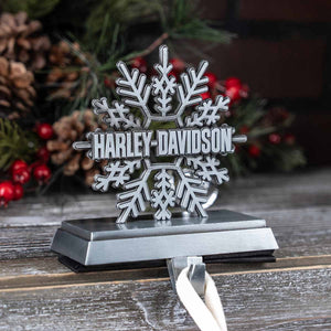 A silver snowflake-shaped Harley-Davidson ornament on a black base with a white ribbon, displayed on a rustic wooden surface with pine and red berries in the background.