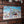 A rectangular metal wall sign featuring a colorful winter holiday illustration, mounted on a red brick wall. The sign depicts Santa Claus, children, and a sculpture in a snowy scene, with the text 'Santa Pole Ice Sculpture Contest' and 'Harley-Davidson' visible on the artwork.