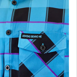 Close-up of a plaid button-up shirt in bright blue, black, and purple, featuring a chest pocket with a black label that reads 'DEMONS BEHIND ME' and matching black buttons.