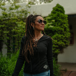 Woman wearing sunglasses and a black top with a logo, standing outdoors with greenery in the background