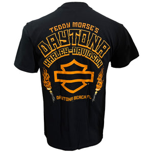Black short-sleeve t-shirt shown from the back, featuring a large orange and yellow graphic print with 'Teddy Morse's Daytona Harley-Davidson' and a flame motif on a white background.